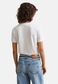 Personne aux cheveux bouclés bruns portant une chemise blanche à manches courtes rentrée dans un jean bleu clair avec une ceinture noire et blanche.