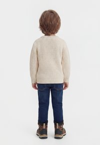 Enfant aux cheveux bruns courts portant un pull en laine crème, un jean bleu et des bottes d'hiver marron, debout de dos sur un fond blanc.