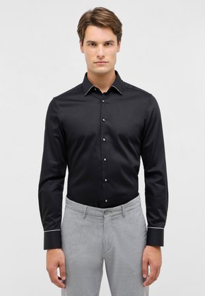 Zwarte langelijfshirt met een gestructureerde kraag, witte accenten, knoopsluiting aan de voorkant en satijnen afwerking, gecombineerd met grijze textuurbroek.