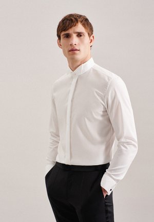 Jeune homme portant une chemise blanche à col mao et un pantalon de costume noir, debout, une main dans la poche, fond neutre.