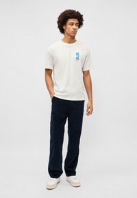 Tricou din bumbac crem cu imprimeu grafic albastru, asortat cu pantaloni din catifea de culoare bleumarin închis și adidași albi; croială lejeră și mâneci scurte.