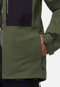 Personne portant une veste vert olive avec la main dans la poche latérale zippée et une couche intérieure noire visible au niveau de la fermeture éclair.