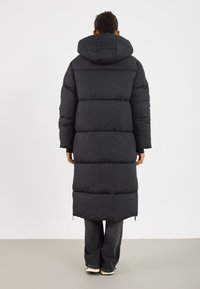 Person iført en lang sort polstret frakke med hætte, som står med ryggen til mod en ensfarvet hvid baggrund.