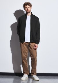 Sort zip-up jakke med tekstureret overdel, båret over en hvid t-shirt, parret med brune bukser og hvide sneakers, mod en ensfarvet baggrund.