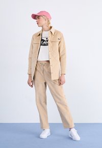 Beige Kord-Jacke mit gestepptem Muster, kombiniert mit beige Kord-Hose und einem weißen, bedruckten Crop-Top. Rosa Baseballkappe und weiße Sneaker.