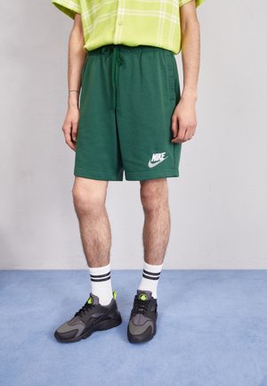 Grønne shorts med elastisk talje og sidelommer, med et hvidt Nike-logo. Båret med sorte og grå sneakers og stribede hvide sokker.