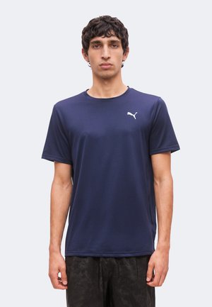 Junger Mann mit kurzen lockigen Haaren, der ein dunkelblaues Puma-Kurzarmshirt und dunkle Hose trägt, steht vor einem schlichten hellen Hintergrund.