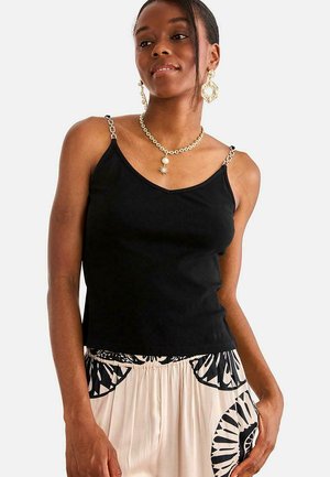 Mujer con top negro sin mangas con tirantes de cadena, falda clara con estampado, pendientes geométricos dorados y un collar dorado con colgante de sol.