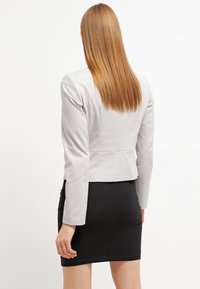 Blazer ajusté gris clair en tissu texturé, manches longues et design court, porté sur une jupe noire ajustée.