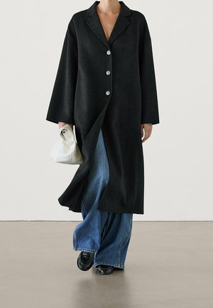Personne portant un long manteau noir boutonné sur un jean bleu à jambes larges, des chaussures noires, et tenant un sac à main blanc plié.