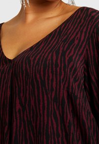 Schwarz-burgunderfarbenes Oberteil mit V-Ausschnitt, weitem Schnitt und gestreiftem Muster. Weiche Textur mit sichtbaren Nähten und kurzen Ärmeln.