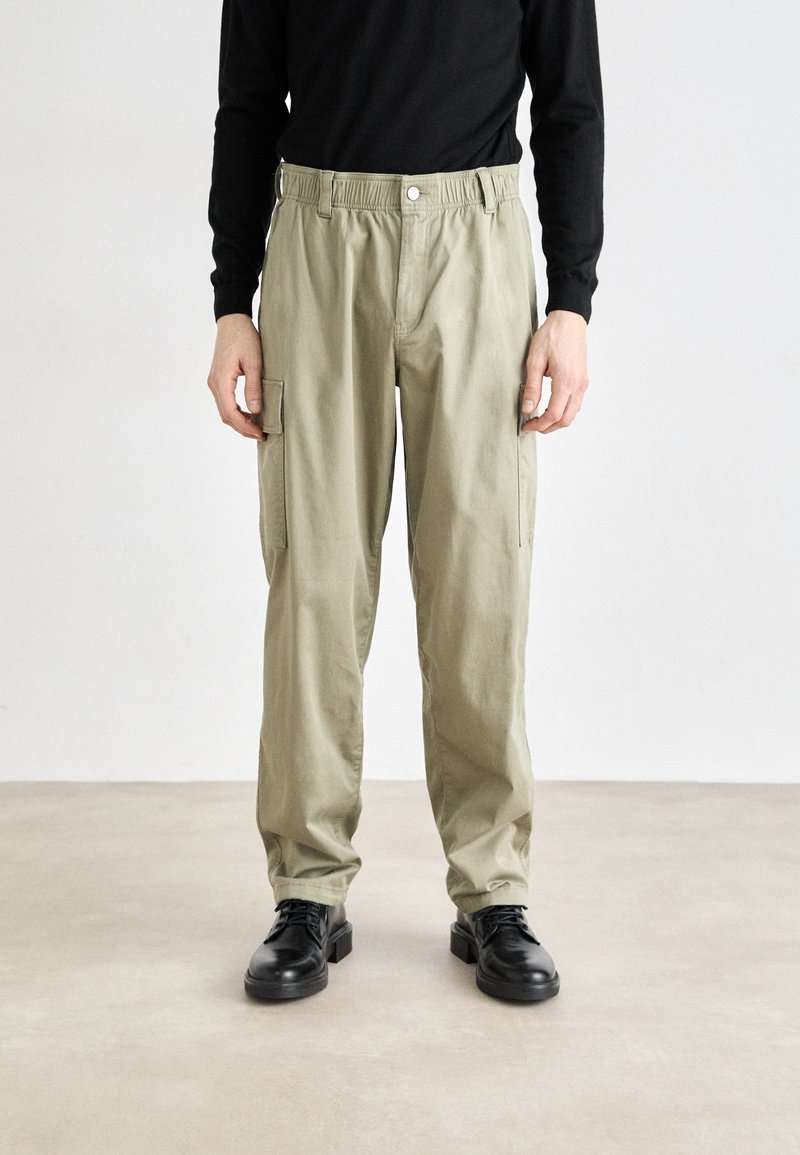 Pantalones cargo caqui con una cinturilla elástica, presentan bolsillos laterales, un ajuste holgado y una tela de algodón suave. Llevados con zapatos de cuero negros.