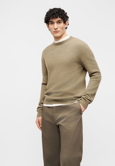 Νέος άνδρας με σγουρά μαλλιά, φοράει μια ανοιχτόκαφέ ζακέτα πάνω από ένα λευκό πουκάμισο και καφέ παντελόνι, στέκεται με το ένα χέρι στην τσέπη.