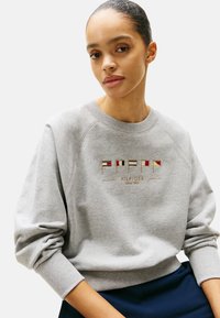 Felpa in cotone grigia con polsini e orlo a costine. Presenta un testo con logo ricamato colorato, insieme a forme geometriche e dettagli di design.