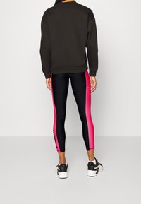 Person, der einen schwarzen Sweatshirt und schwarze Leggings mit breiten rosa Seitenstreifen sowie schwarz-weißen Sneakers trägt, steht vor einem einfarbigen Hintergrund.