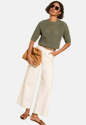 Olivgrüner gestrickter Pullover mit kurzen Ärmeln, kombiniert mit cremefarbenen weit geschnittenen Hosen, hält eine tannfarbene Wildleder-Clutch und trägt braune Slide-Sandalen.