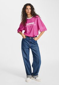 Rosa t-shirt med kort ärm med "hummel PERFORMANCE"-logotyp, i kombination med lösa blå jeans och vita sneakers med mörka detaljer. Slät textur.