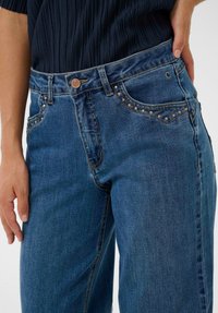 Blå denimjeans med højtaljet design, udstyret med kobberbeslag og nitter på lommerne. Bukserne har lige ben og en mellemblå vask.