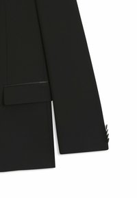 Primo piano di una manica di giacca nera con quattro bottoni e taschino a patta, realizzata in tessuto leggermente strutturato, su sfondo bianco.