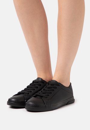 Fødder iført sorte lavtop snørestøvler med glat tekstur på hvid baggrund.