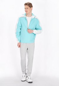 Giacca a vento azzurra chiara con maniche bianche, frontale con zip e cappuccio regolabile, abbinata a pantaloni della tuta grigi e sneakers bianche.
