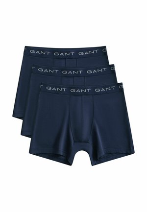 Trois caleçons pour hommes bleu marine avec logo GANT sur les ceintures élastiques, empilés en chevauchement sur un fond blanc.