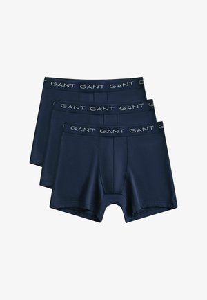 Trois caleçons pour hommes bleu marine avec logo GANT sur les ceintures élastiques, empilés en chevauchement sur un fond blanc.