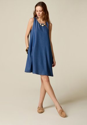 Mujer con vestido vaquero azul sin mangas, collar y pulsera, de pie con las piernas cruzadas y zapatos tipo mocasín color crema.