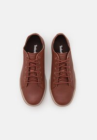 Pár hnědých kožených tenisek Timberland s béžovými podrážkami a tkaničkami, z pohledu shora na bílém pozadí.