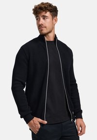 Musta vetoketjullinen takki, jossa on teksturoitu neulospatterni, resorit hihansuissa ja valkoinen kontrastivetoketju. Päällä musta t-paita.