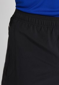 Svarta träningsshorts med elastisk midjeband, slät textur och sidofickor synliga i bilden. Tyget verkar lätt.