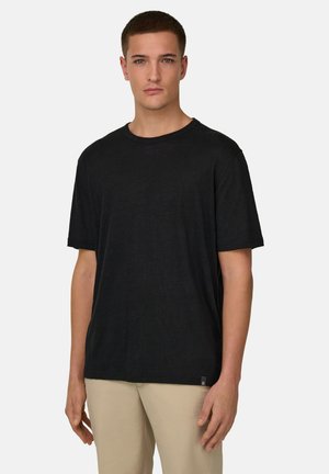 Giovane uomo con capelli corti che indossa una t-shirt nera semplice e pantaloni beige, in piedi davanti a uno sfondo chiaro e uniforme.