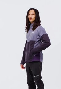 Lilla og grå pullover med høj krave og sidelommer, parret med sorte bukser med et 'TERREX' logo på benet.
