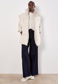 Gewatteerde mouwloze vest in lichtbeige, met een hoge kraag, zijzakken en een zachte textuur. Gemaakt over een gestructureerde witte trui en donkere jeans.