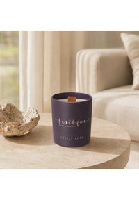 Bougie violette étiquetée velvet rose avec mèche en bois sur une table ronde en pierre à côté d'un bol décoratif et d'un canapé beige en arrière-plan.