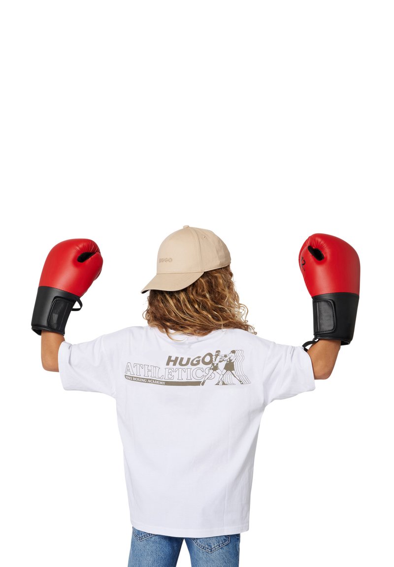 Gants de boxe rouges, t-shirt blanc avec un graphique "HUGO ATHLETICS", casquette beige claire et shorts en denim. Vue arrière du porteur.