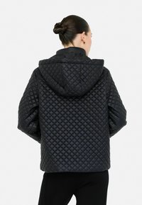 Vrouw met zwart haar in een knot, gekleed in een zwarte gewatteerde jas met capuchon, van achteren getoond tegen een effen witte achtergrond.