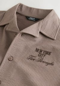 Brun, struktureret skjorte med krave. Har "NEW YORK CITY" og "Fire Boroughs" broderet i mørkebrun.