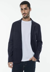 Mörk marinblå blazer med en slät textur, snedskurna slag, enkel knappstängning och framfickor över en vit t-shirt och mörka jeans.