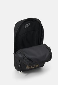 Zaino nero con una finitura liscia, chiusura con zip e tasca piatta. L'interno è foderato in tessuto nero; logo visibile all'interno.