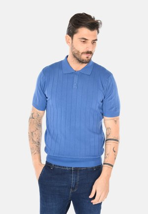 Uomo con capelli corti e scuri che indossa una polo a maniche corte blu a coste e jeans scuri, con le mani nelle tasche, guarda verso il basso e a destra.
