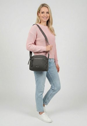 Frau lächelt, trägt einen rosa Pullover, hellblaue Jeans, weiße Sneaker und eine schwarze Umhängetasche über der Schulter.