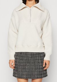 Bílé fleecové pulovry s ribbed límcem a polovičním zipem, v kombinaci s texturovanou sukní z černobílé tkaniny. Útulný a moderní design.