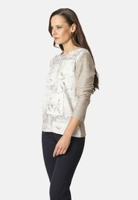 Una mujer con cabello largo y castaño, vestida con una blusa de manga larga beige con un patrón y pantalones oscuros, de perfil frente a un fondo liso.