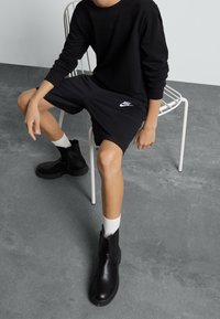 Černá mikina a šortky s bílým logem Nike, doplněné černými kotníkovými botami a bílými ponožkami, sedící na bílé kovové židli.