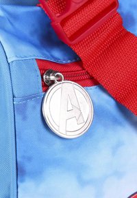 Blue fabric bag with red strap and zipper. Features a silver circular tag with an engraved "A" logo. Textured surface and bold colors.