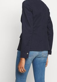 Veste bleu marine, design ajusté avec manches longues. Comprend un ourlet arrondi et des boutons argentés en décoration sur les poignets. Tissu texturé.