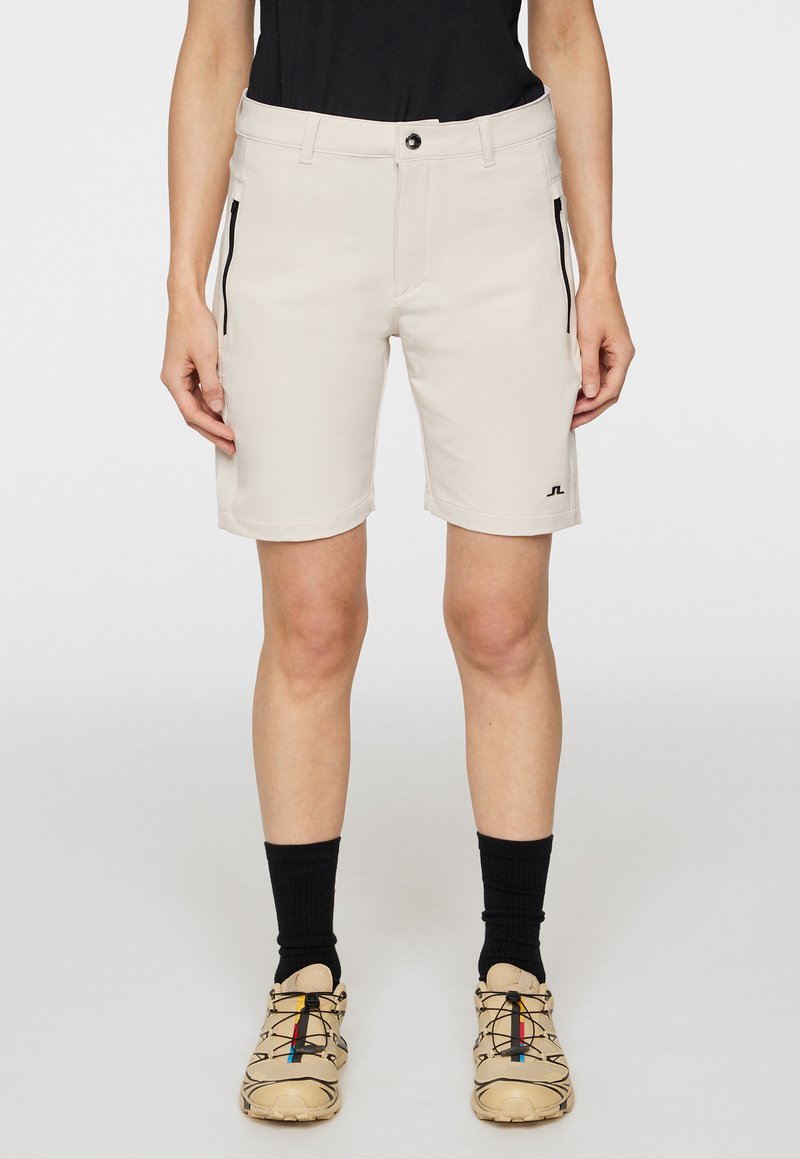 Beiga shorts med en slät textur, bältesömmar och två zipparade sidofickor. Har en logotyp på nedre benet, i kombination med beiga skor och svarta strumpor.