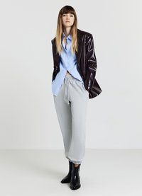 Donna in posa con le mani nelle tasche, indossando una camicia azzurra a bottoni, una giacca scura lucida, pantaloni jogger grigi e stivali alla caviglia neri su uno sfondo neutro.