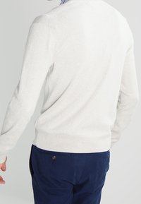 Pull en maille gris clair avec un col rond, des poignets et un ourlet côtelés, porté sur un pantalon bleu marine, montrant un tissu lisse et texturé.